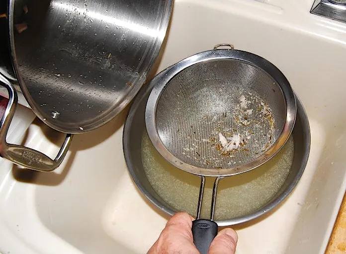 傾けた鍋の下で金属のざるを手で支え、ざるの中に香草や肉のかすが残り、下の鍋に漉した煮汁がたまっている