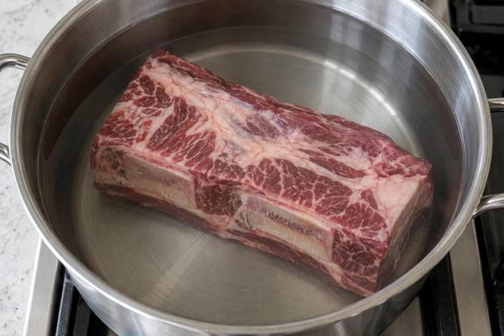 深鍋の透明な水に沈めた、ひと塊の生の骨付き牛バラ肉